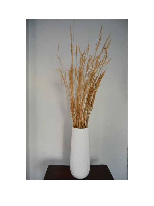 Dried Grass bunch in a white medium tall vase on top of a brown stool. Background is white. 