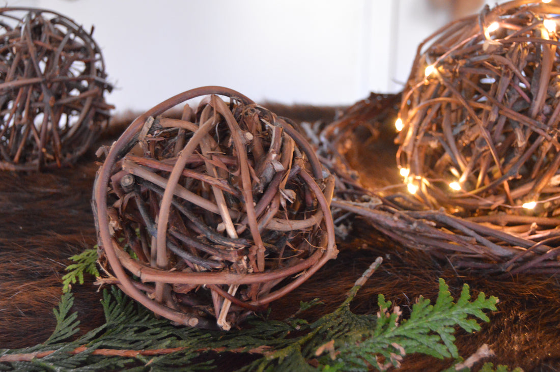 Three balls made of grapevine. One of those balls are wrapped in small fairy lights, placed in the middle of a grapevine circle. Balls are placed on top of evergreen pieces and placed on top of brown fur.